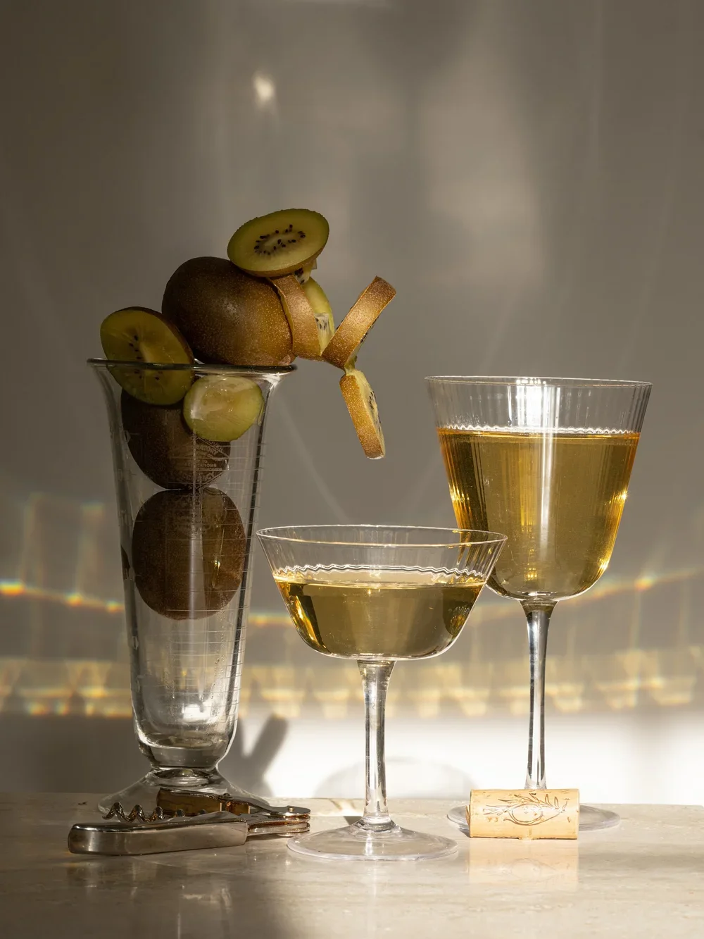 A clear tall glass with kiwi fruit slices and a whole kiwi on top, a smaller glass with a yellow drink, a cocktail spoon, and a glass of whiskey on a light-colored surface with sunlight reflections.