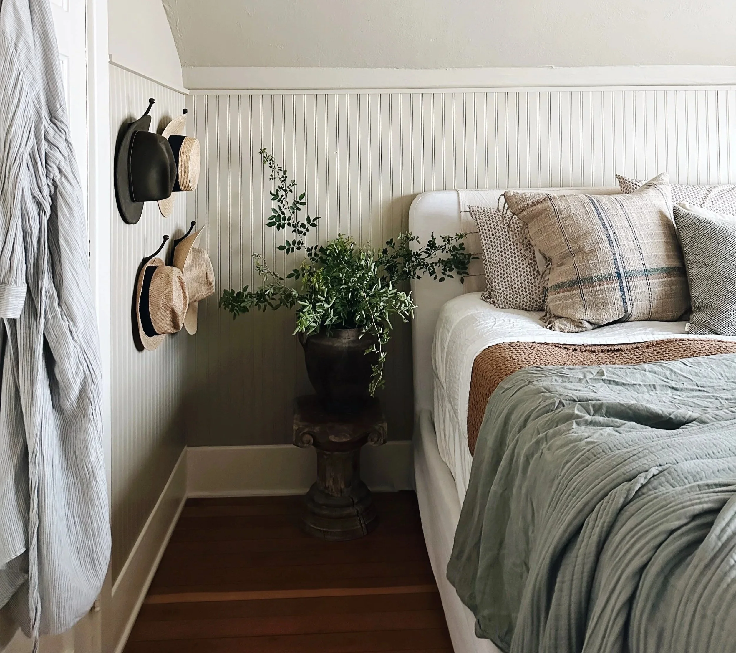 Cozy bedroom corner with a bed dressed in plaid and solid colored pillows, a large potted plant on an antique wooden stand, and hats hanging on the wall with a striped garment hanging on the side.