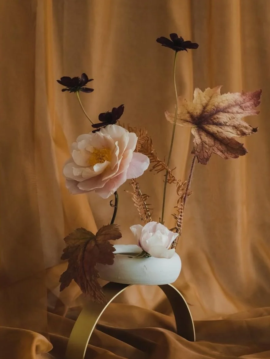 A decorative floral arrangement in a white ceramic vase with black and white flowers, dried leaves, and ferns, all on a curved golden stand against a brown curtain background.