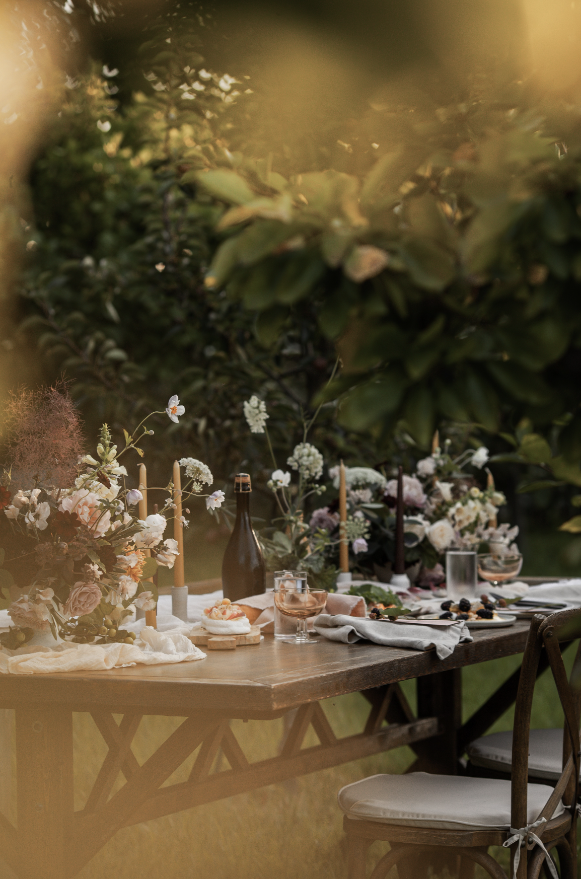 A rustic outdoor dining table decorated with flowers, candles, and tableware, set in a lush garden with trees and greenery in the background.