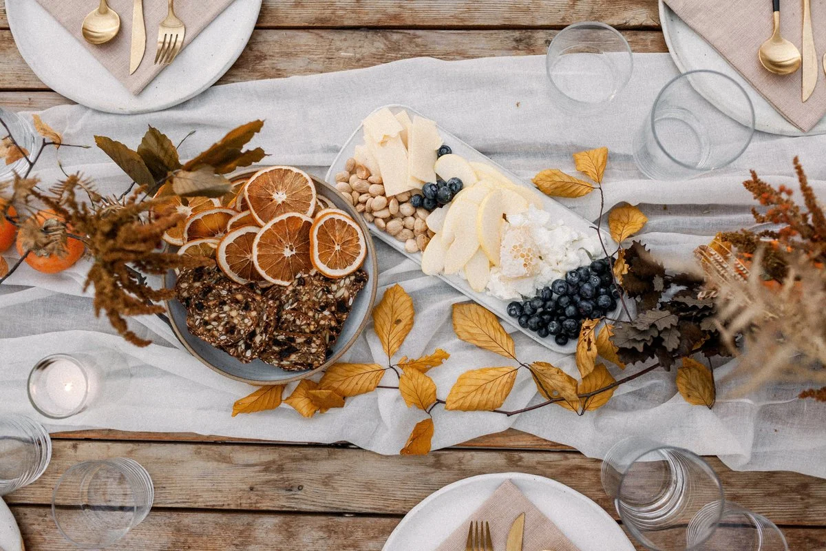A fall-themed table centerpiece with dried leaves, a cheese platter with grapes and nuts, dried orange slices, and chocolate bark with nuts, surrounded by plates, gold utensils, and empty glasses on a wooden table.