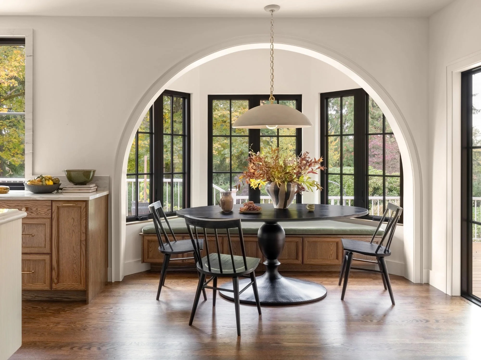 Dining area with round black table, four black chairs, a built-in window seat with cushions, and large windows showing outside greenery.