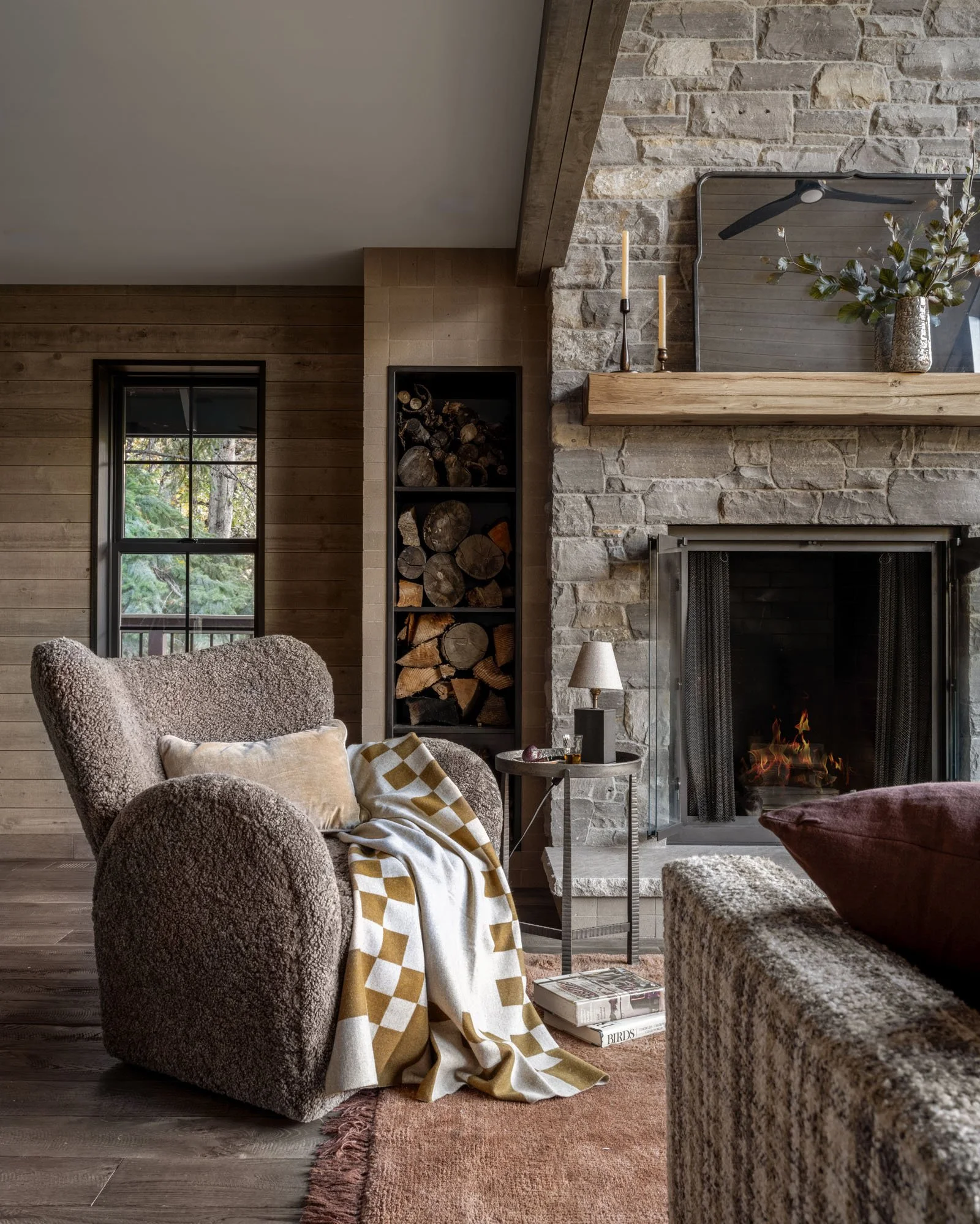 Living room with a cozy armchair, a fireplace, and logs stored in a built-in nook, natural light coming through a window, and decorative items on the mantel.