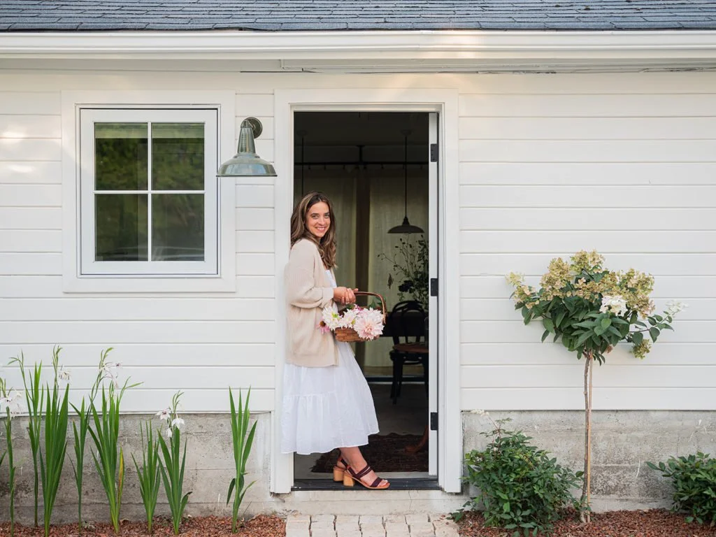 A woman is standing at the doorway of a white house, holding a basket of flowers, wearing a white skirt and beige sweater, smiling, with a garden and plants outside.
