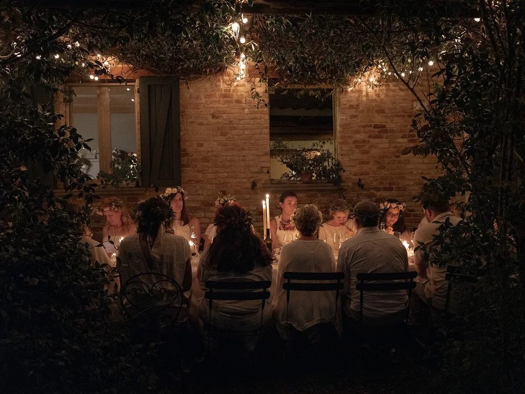 People sitting around a dinner table during a candlelit gathering in an outdoor setting at night, surrounded by foliage and string lights.