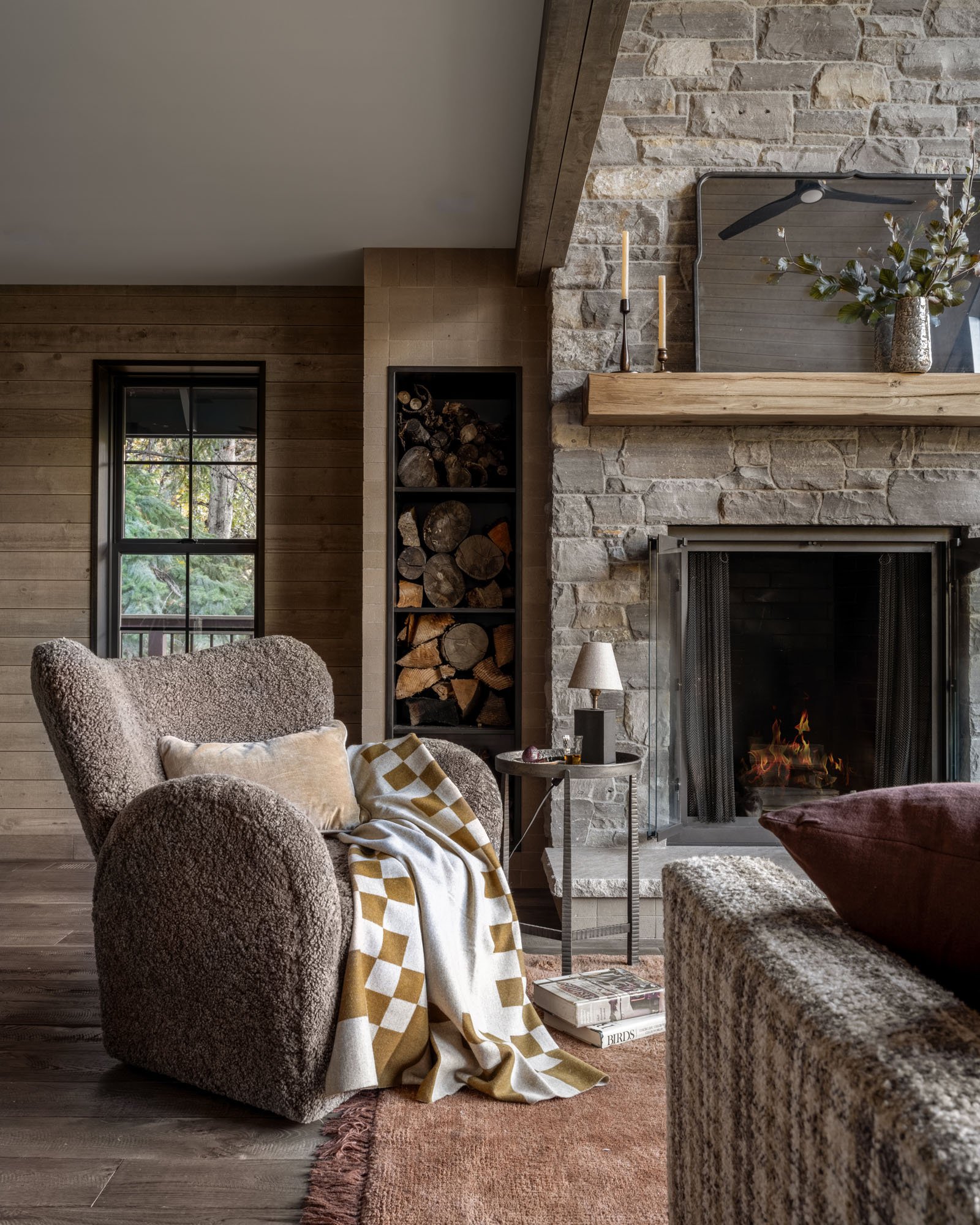 Cozy living room with a stone fireplace, wood-paneled wall, large armchair with a beige pillow and throw, side table with lamp, wood logs stored in an inset wall, and a window with trees outside.