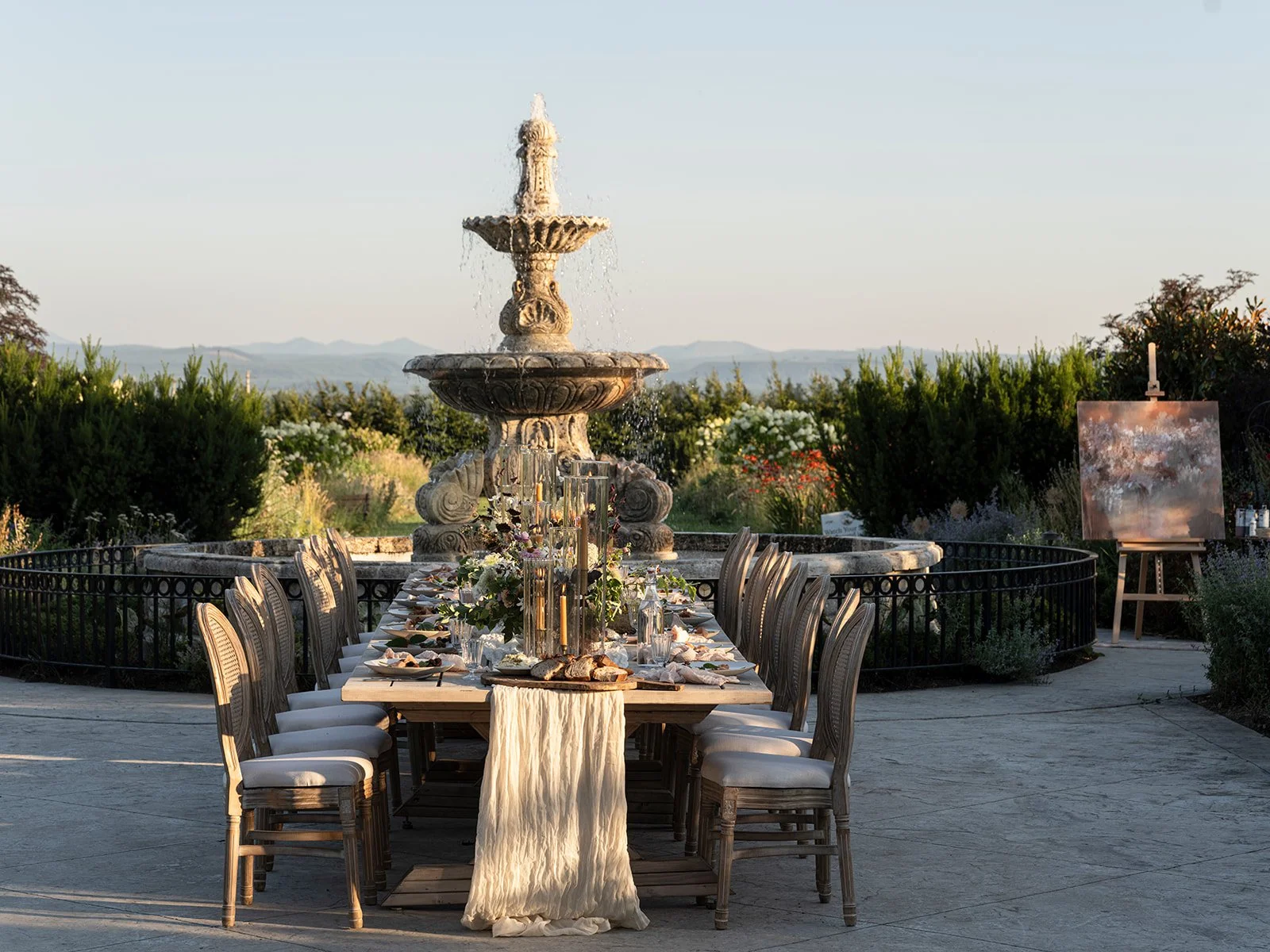 Outdoor dining table set for a meal with a fountain in the background surrounded by greenery, a painting on an easel, and mountain views in the distance.