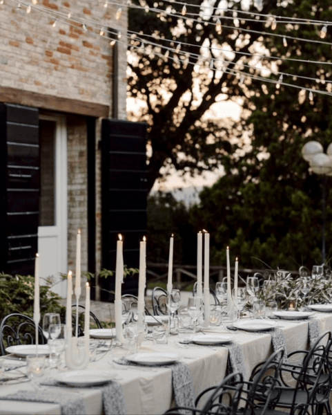 Long outdoor dining table set with white tablecloths, tall white candles, glassware, and chairs, during sunset with string lights overhead and trees in the background.