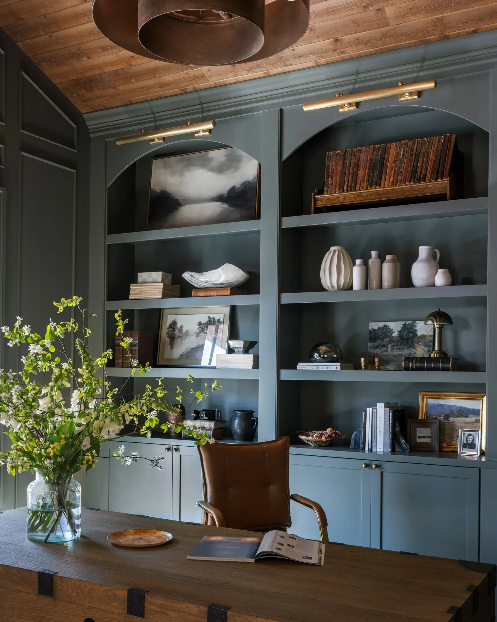 A room with a wooden ceiling, dark green built-in shelves with decorative items and books, and a wooden table with a glass vase of white and green flowers, along with an open magazine.