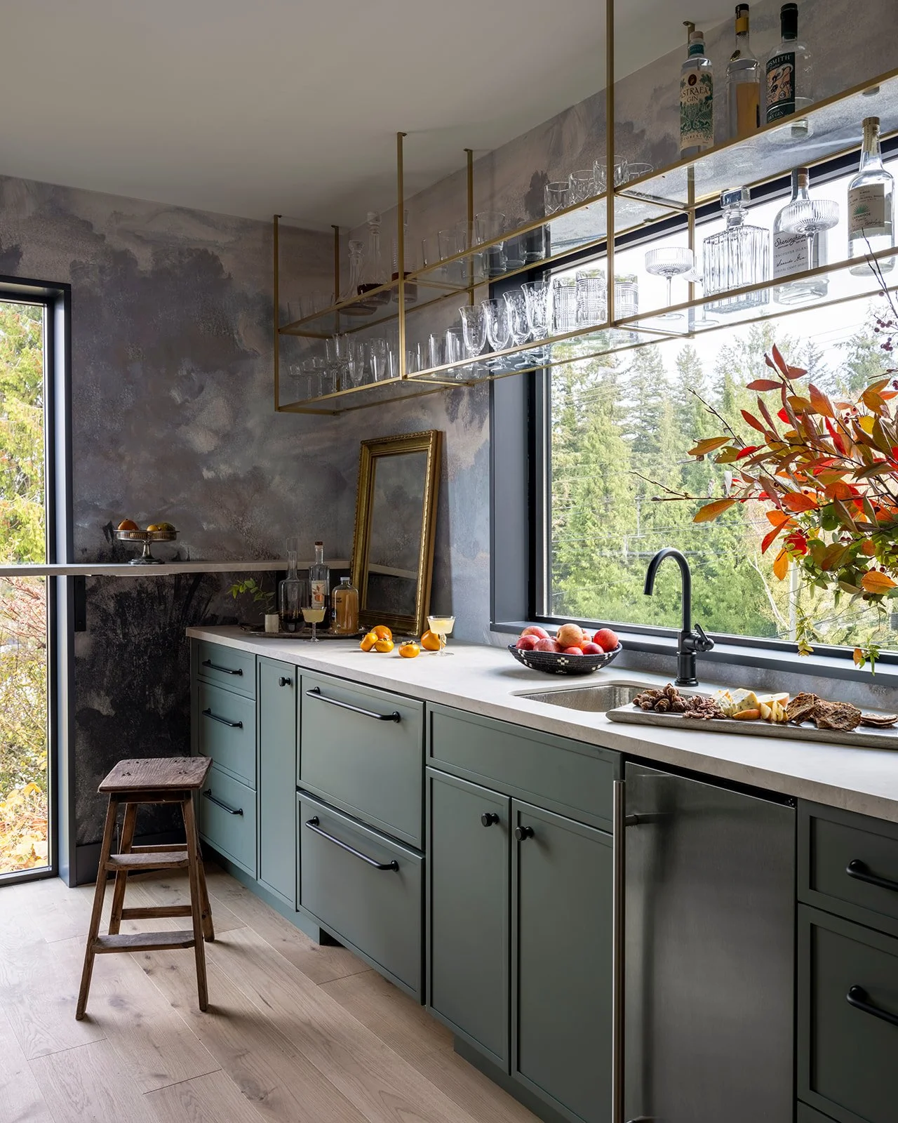 Modern kitchen with green cabinetry, a marble countertop, open shelving with glassware, a window overlooking trees, and a wooden stool.