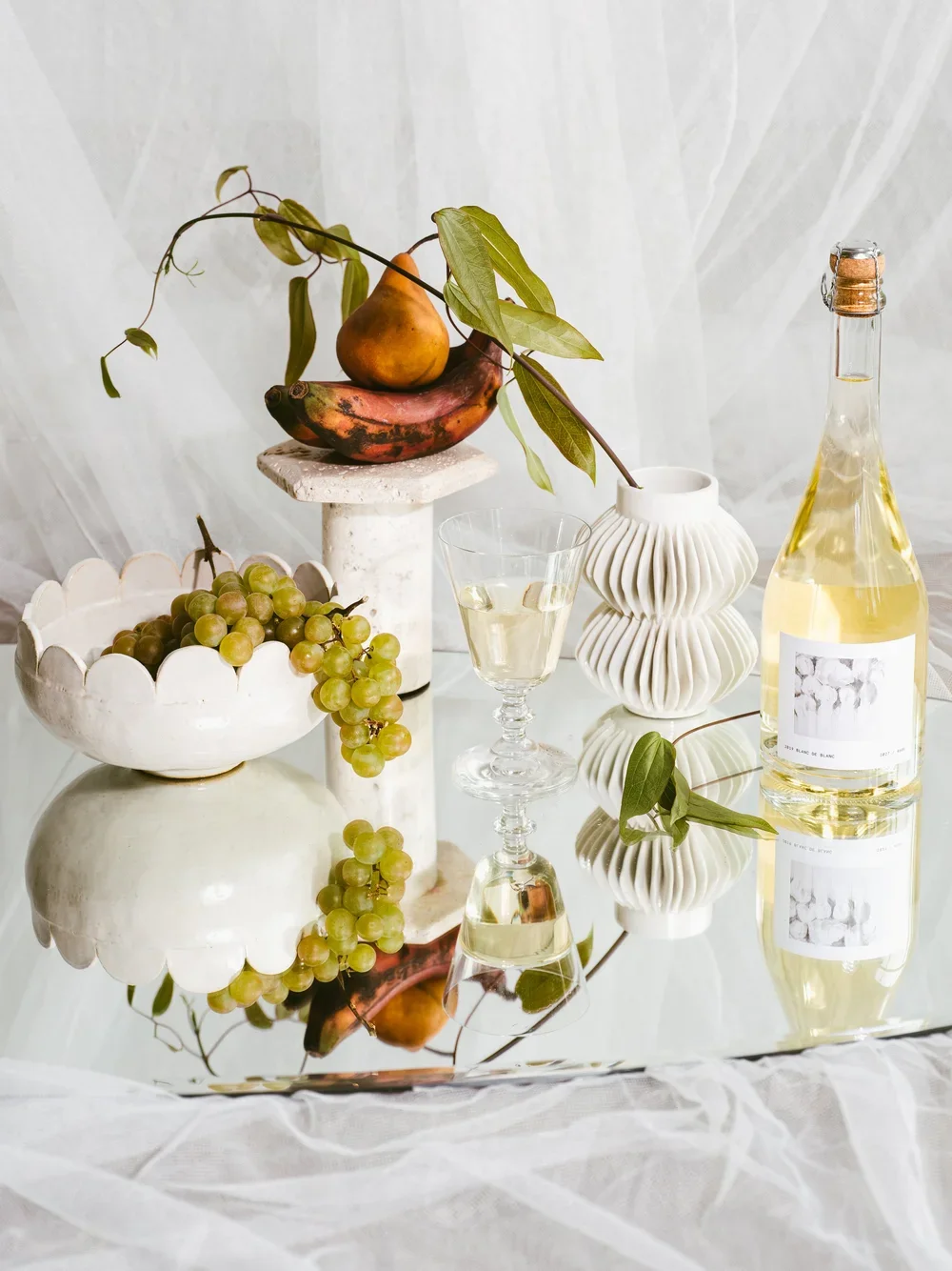 A still life arrangement with grapes, a pear, and a banana on a pedestal, a glass of white wine, a decorative white vase, a wine bottle, and a white bowl with grapes, on a reflective surface with a white background.