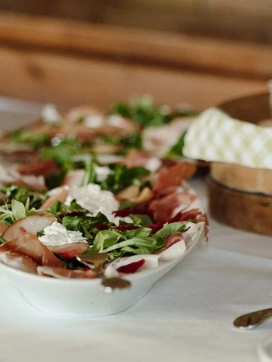 A bowl of salad with leafy greens, sliced meat, and crumbled white cheese on a white surface, with a wooden cutting board and a wrapped cloth in the background.
