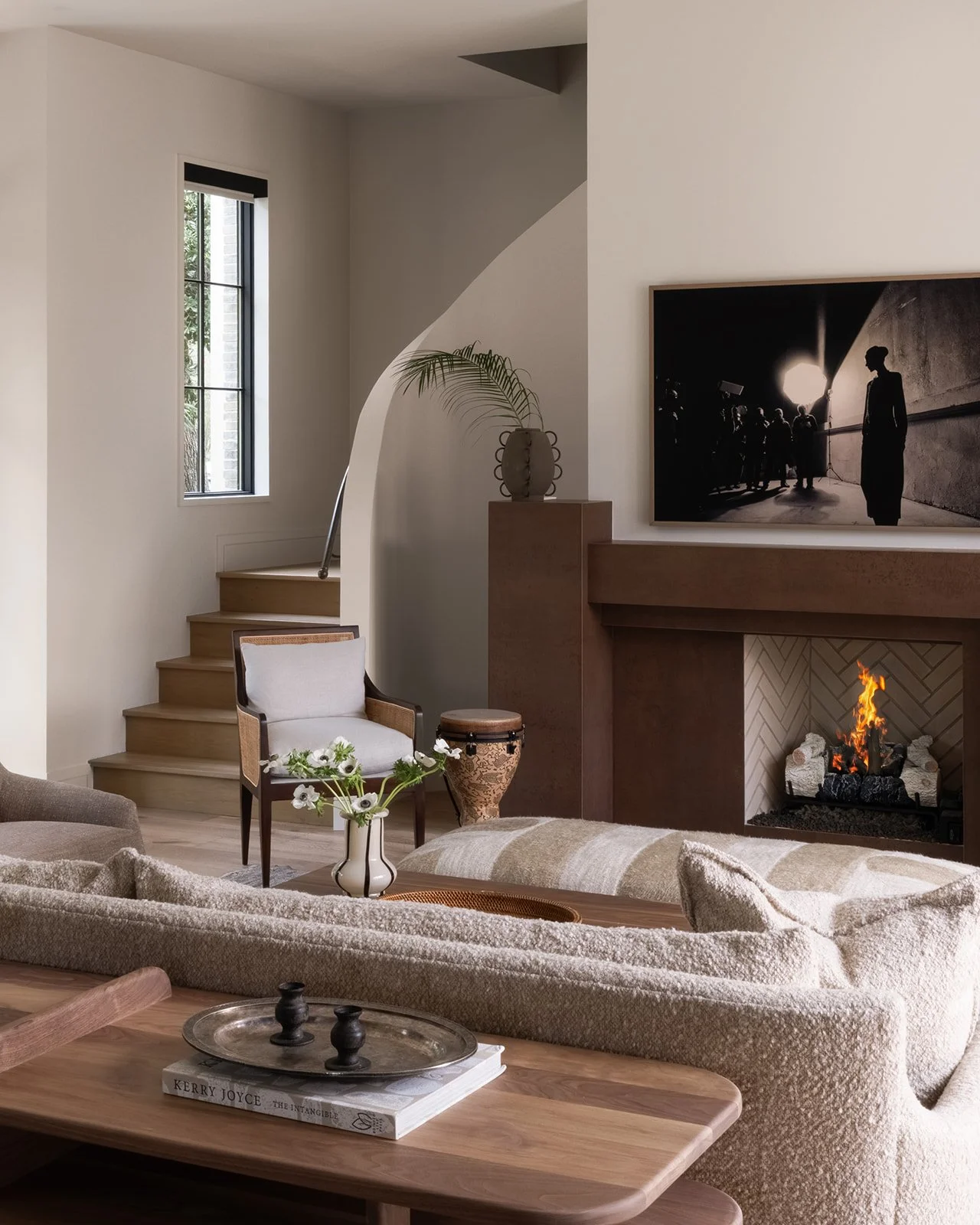 Living room with fireplace, black and white artwork, armchair, flowers, and staircase.
