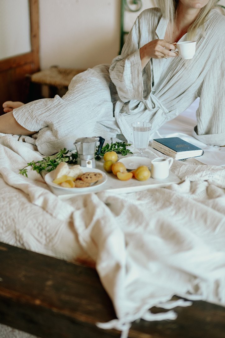 Woman in a bathrobe sitting on a bed, holding a cup, with breakfast items and a book on the bed.