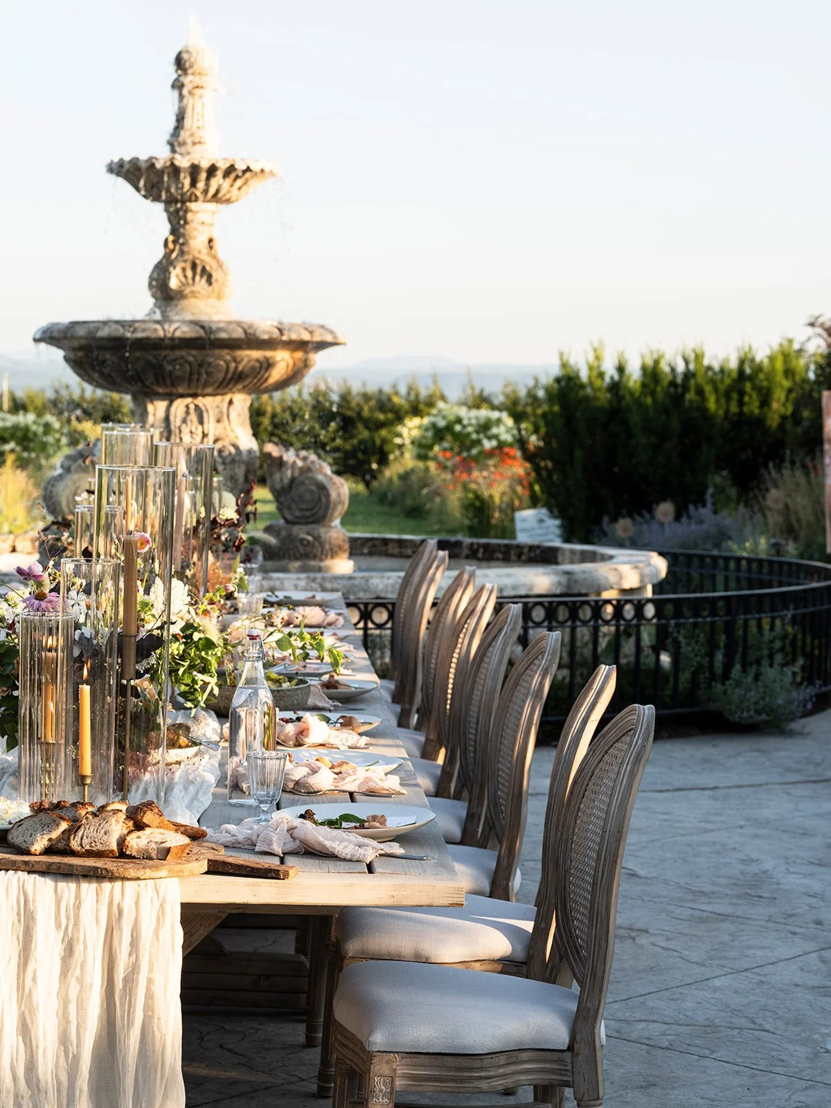 Outdoor banquet table set with plates, glasses, bread, floral centerpieces, and tall candle holders, with a stone fountain and garden in the background.