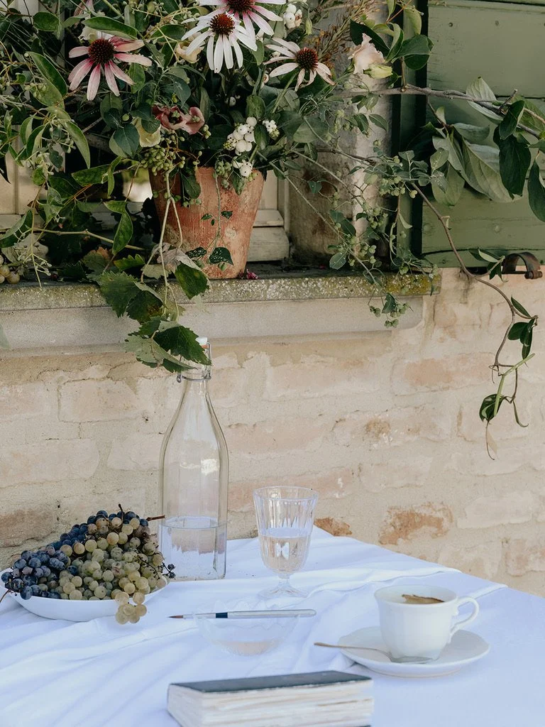 A table with a white tablecloth, grape bunches, a glass of water, a cup of coffee, a pen, a closed book, and a glass bottle, set outdoors against a brick wall and flower arrangement.
