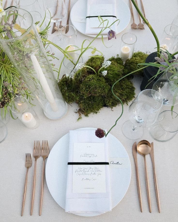 Elegant table setting with white plate, gold cutlery, white napkin, and handwritten menu card, centerpiece with moss and white flowers, candles, and glassware.