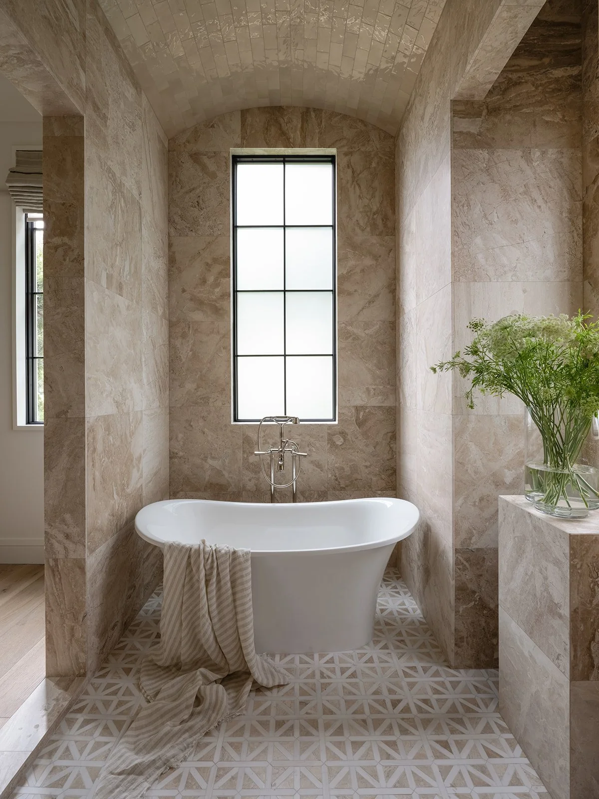 A modern bathroom with a freestanding white bathtub positioned in front of a large window with frosted glass and black grid. The walls and arch are covered with beige marble tiles, and a patterned tile floor. A glass vase with green foliage is on a ledge to the right of the tub, and a beige towel hangs over the edge of the bathtub.