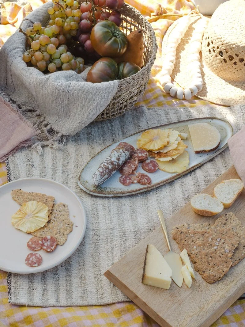 A picnic scene with a basket of grapes, tomatoes, crackers, cheese, and sliced bread arranged on a striped blanket.