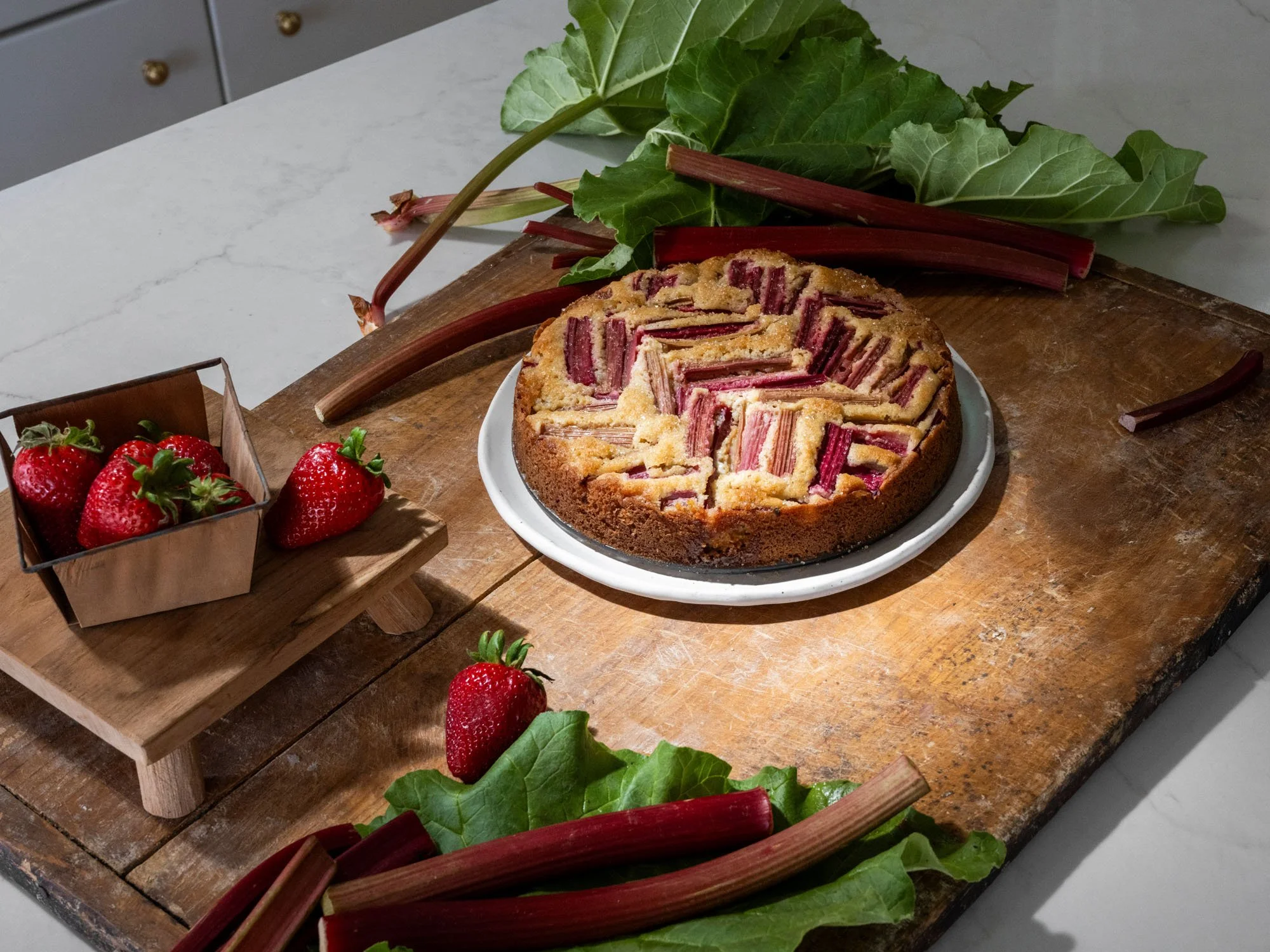 Rhubarb strawberry cake on a white plate, fresh strawberries in a small wooden box, and rhubarb stalks with leaves on a rustic wooden cutting board.