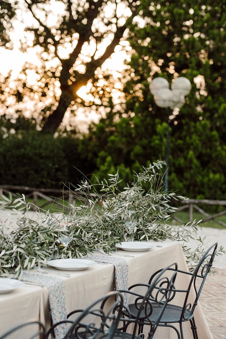 Outdoor dining table set with white plates, glasses, and a long leafy green centerpiece, with a sunset or sunrise in the background and a decorative Mickey Mouse-shaped balloon.