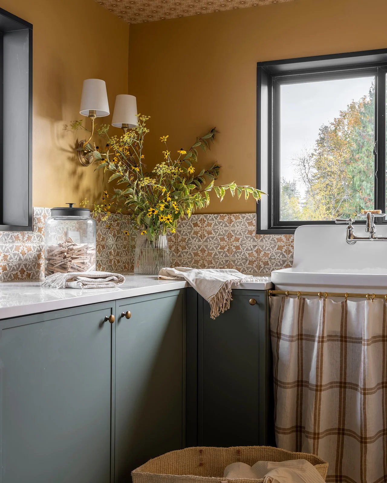 A cozy kitchen corner with a green cabinet, a white marble countertop, a window with a view of trees, and a large glass jar with wooden clothespins. There are decorative towels, a vase with yellow flowers and green foliage, and a wall-mounted lamp with two white shades.