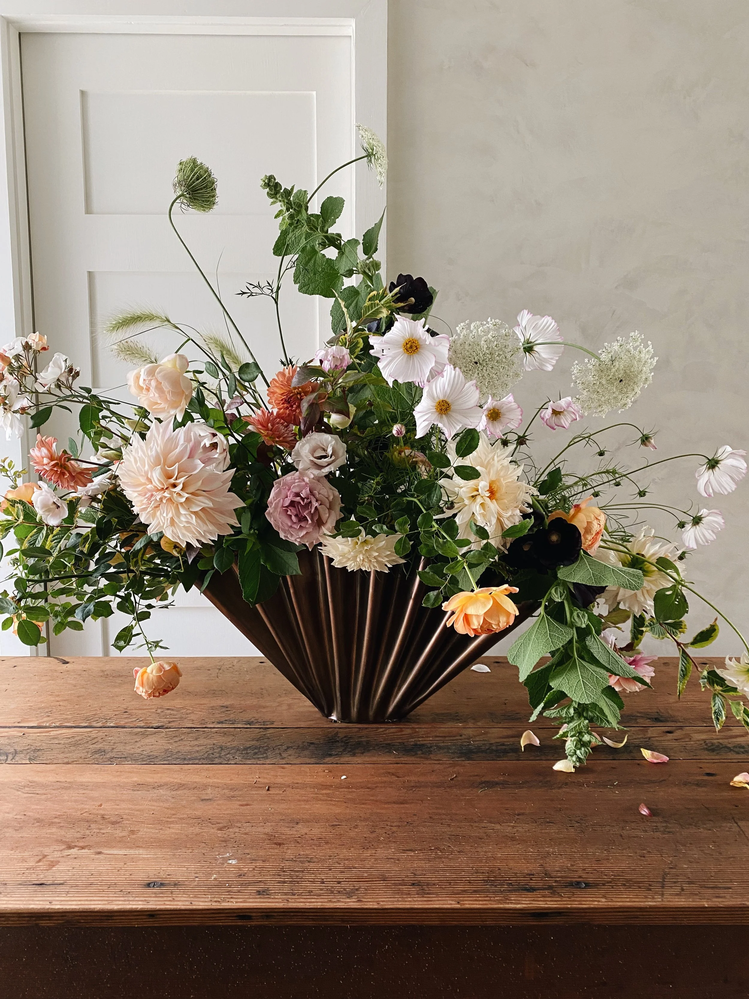 A large bouquet of mixed flowers in a dark, ribbed metal vase on a wooden table in front of a white door and neutral-colored wall.