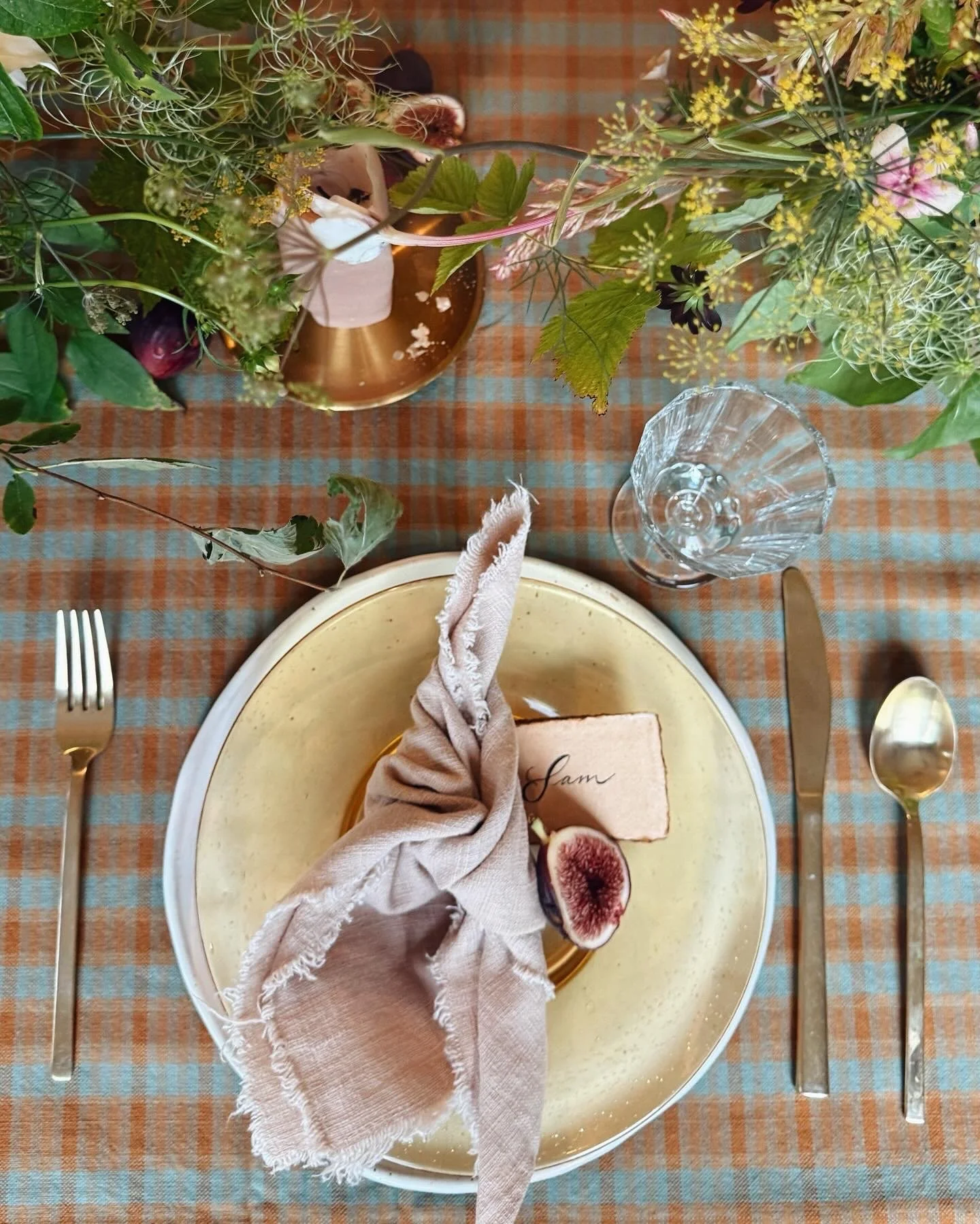 A place setting with a beige napkin folded on a round beige plate, a slice of bread with a fig and a note reading 'Jam', a fork on the left, a knife on the right, and a gold spoon on the right, all on a checked orange and blue tablecloth. There is a glass of water and a floral centerpiece with various flowers and greenery in the background.