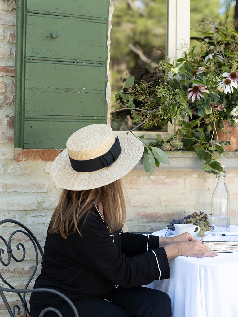 A woman with light brown hair wearing a wide-brimmed straw hat with a black ribbon, sitting outdoors at a table with a white tablecloth, near a brick house with green wooden shutters and a flower box with pink and white flowers, garden scene in background.