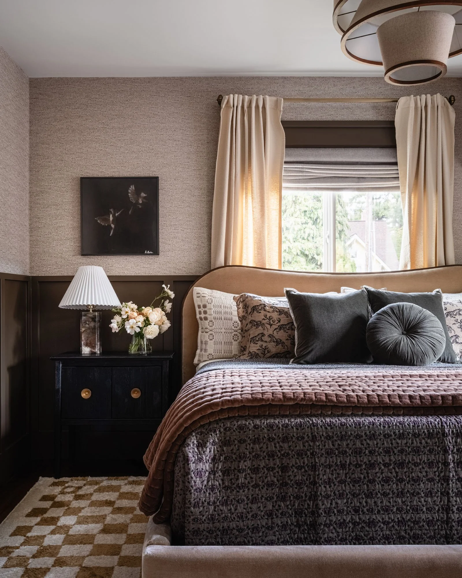 A cozy bedroom with beige curtains, a window, a bed with a plush headboard, multiple decorative pillows, a bedside table with a lamp, a flower vase, a framed art piece on the wall, and a checkered rug.