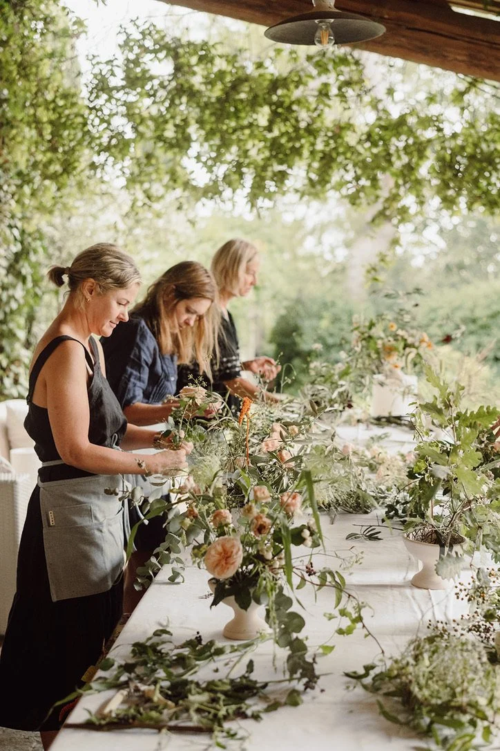 Three women create floral arrangements on a long outdoor table decorated with flowers and greenery, under a wooden roof with string lights, surrounded by trees.