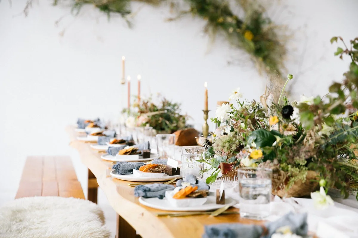 A long wooden dining table set with white plates, glasses, linens, and candles, decorated with an arrangement of flowers and greenery.