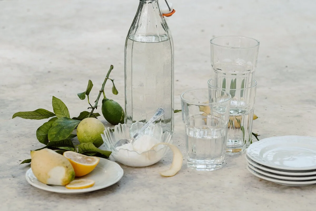 A still life of a glass bottle of water, four glasses filled with water, a plate with sliced lemon, lime, and apple, a bowl of yogurt or sour cream with a spoon, and a stack of small plates on a marble counter with lime and lemon leaves and a lemon and lime still on the counter.