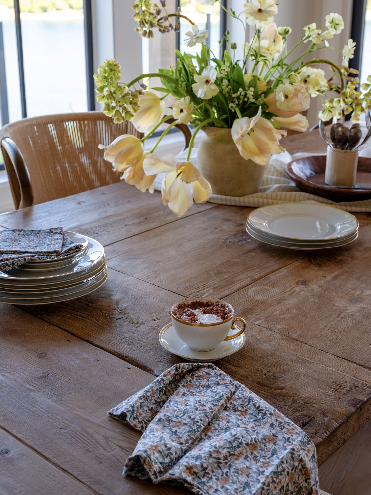 A wooden dining table with a vase of white and pale pink flowers, a stack of white plates, a cup of coffee or hot chocolate, a cloth napkin, and a small container with utensils near a window.