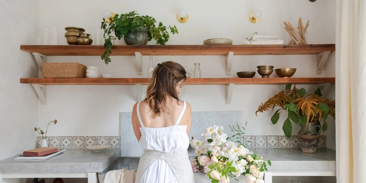 Woman with shoulder-length brown hair arranging a bouquet of white and pink flowers in a kitchen or floral shop with open wooden shelves, plants, and decorative bowls.