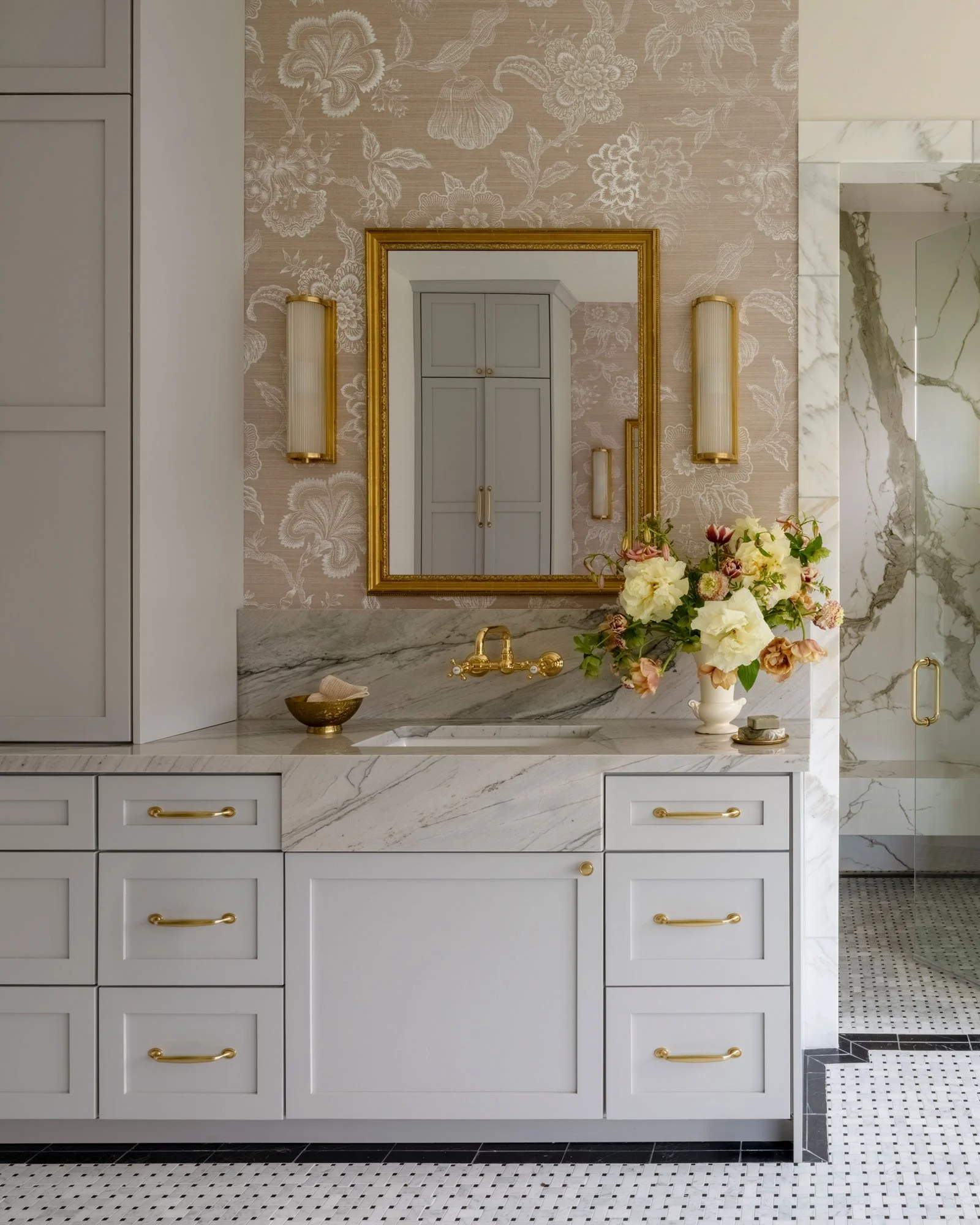 Elegant bathroom with white cabinetry, marble countertop, gold fixtures, a large mirror, floral wallpaper, a vase of flowers, and a glass shower with marble tiles.