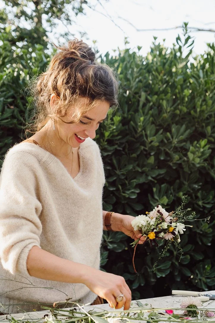 A woman with curly hair smiling while making a flower bouquet outdoors.