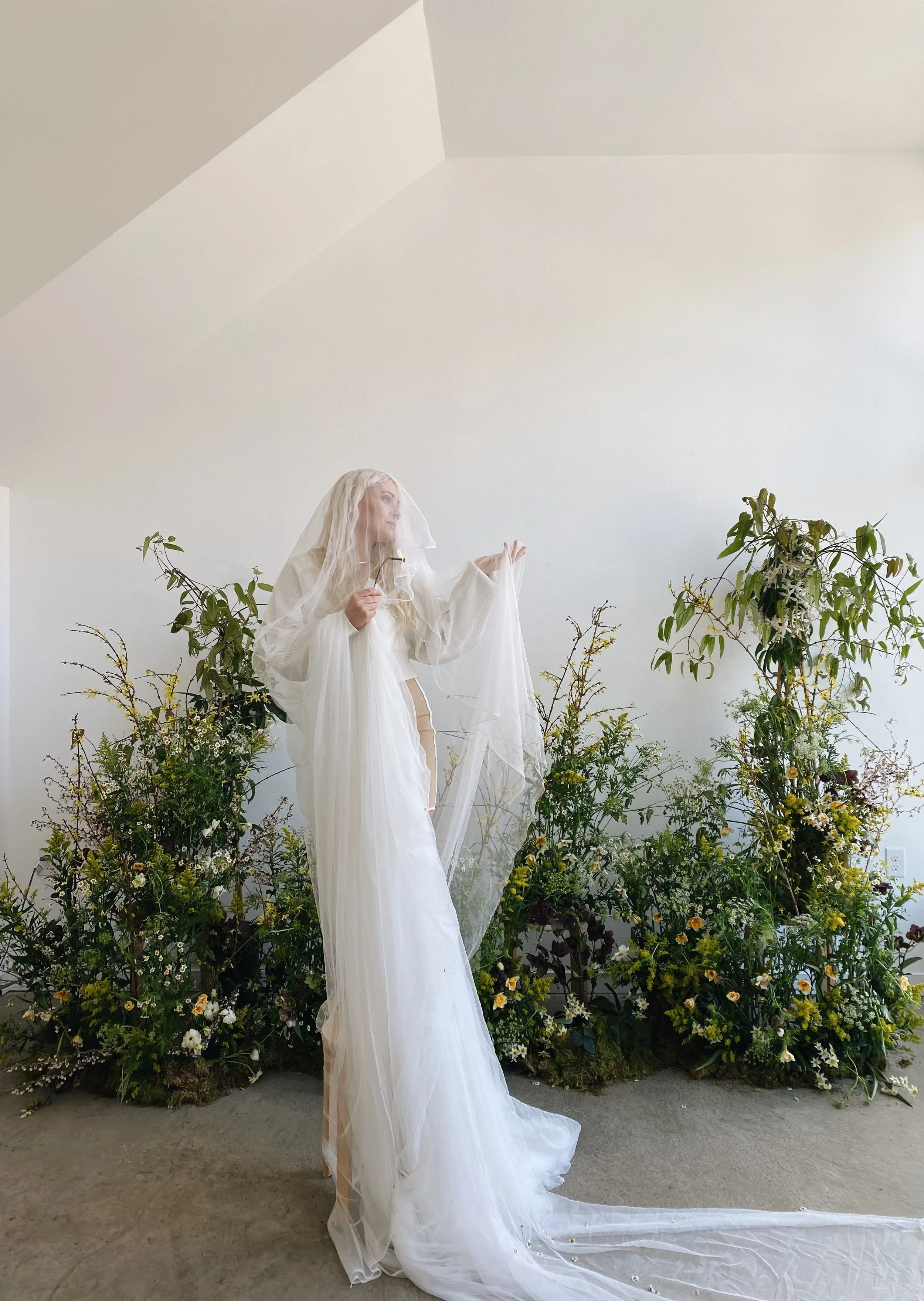 A woman dressed in white with a veil standing among greenery and flowers, holding a piece of fabric and a lipstick, in a minimal modern indoor setting.