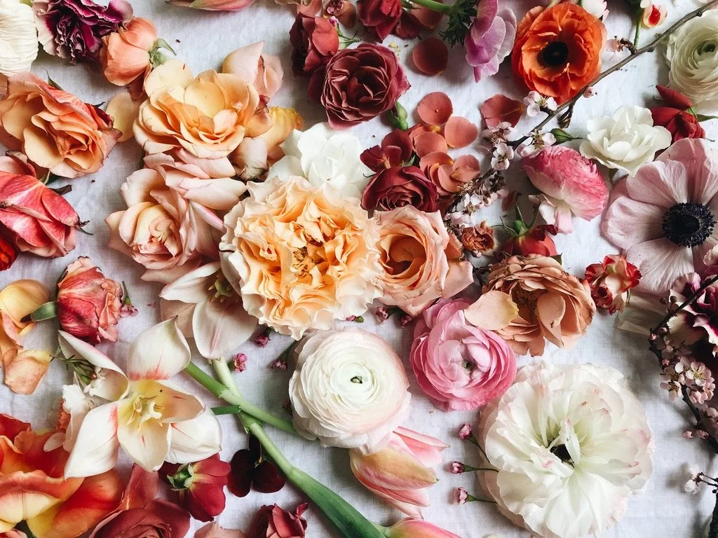 A variety of colorful flowers including roses, ranunculus, anemones, and other blooms arranged on a light-colored surface.