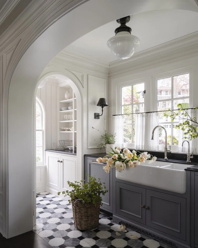 Bright kitchen with white walls and black countertops, farmhouse sink with flowers, small potted plant in basket, patterned tile floor, large windows, and built-in shelves.