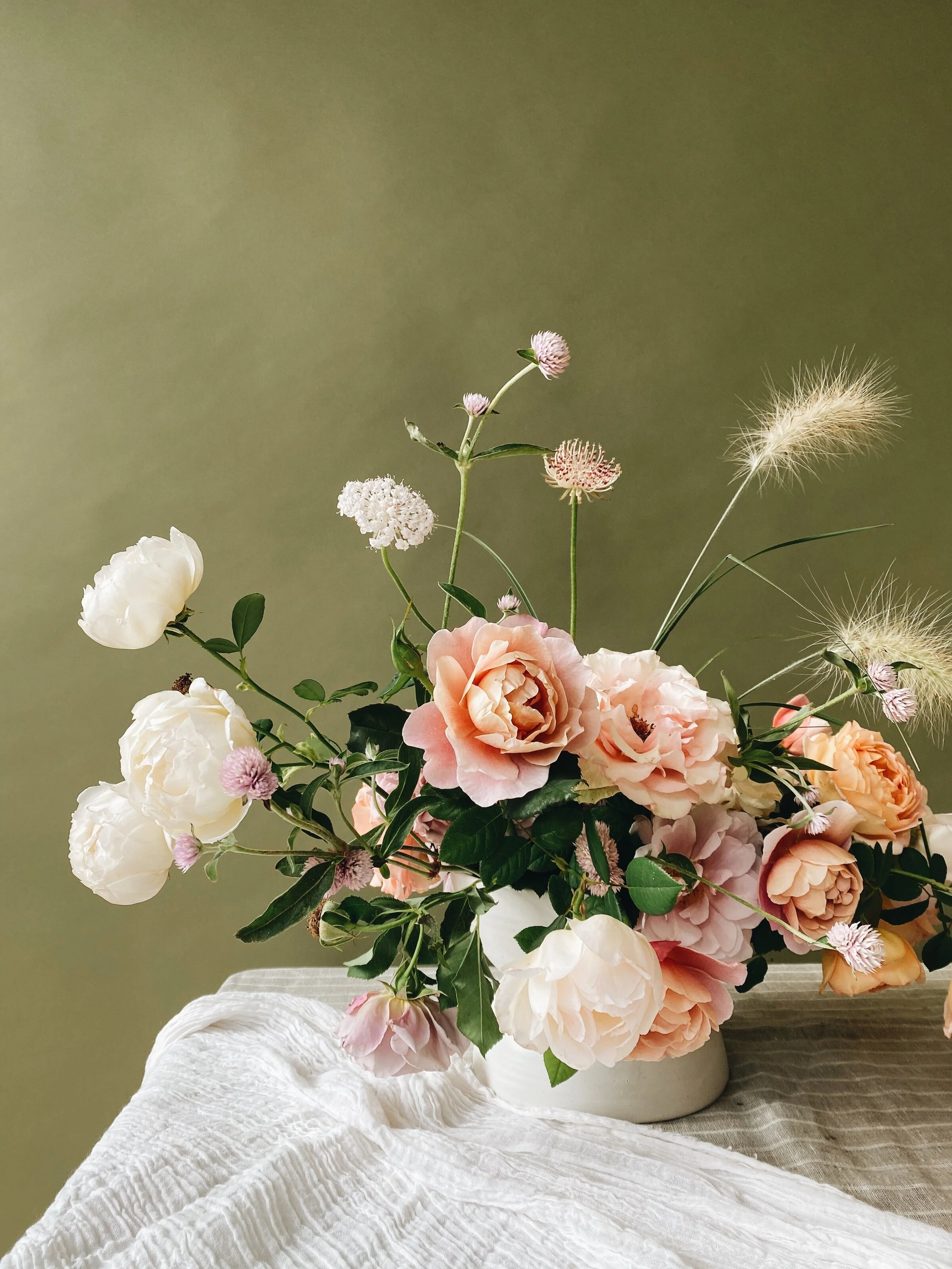 A flower arrangement with pink and white flowers and green leaves in a white vase on a white tablecloth.
