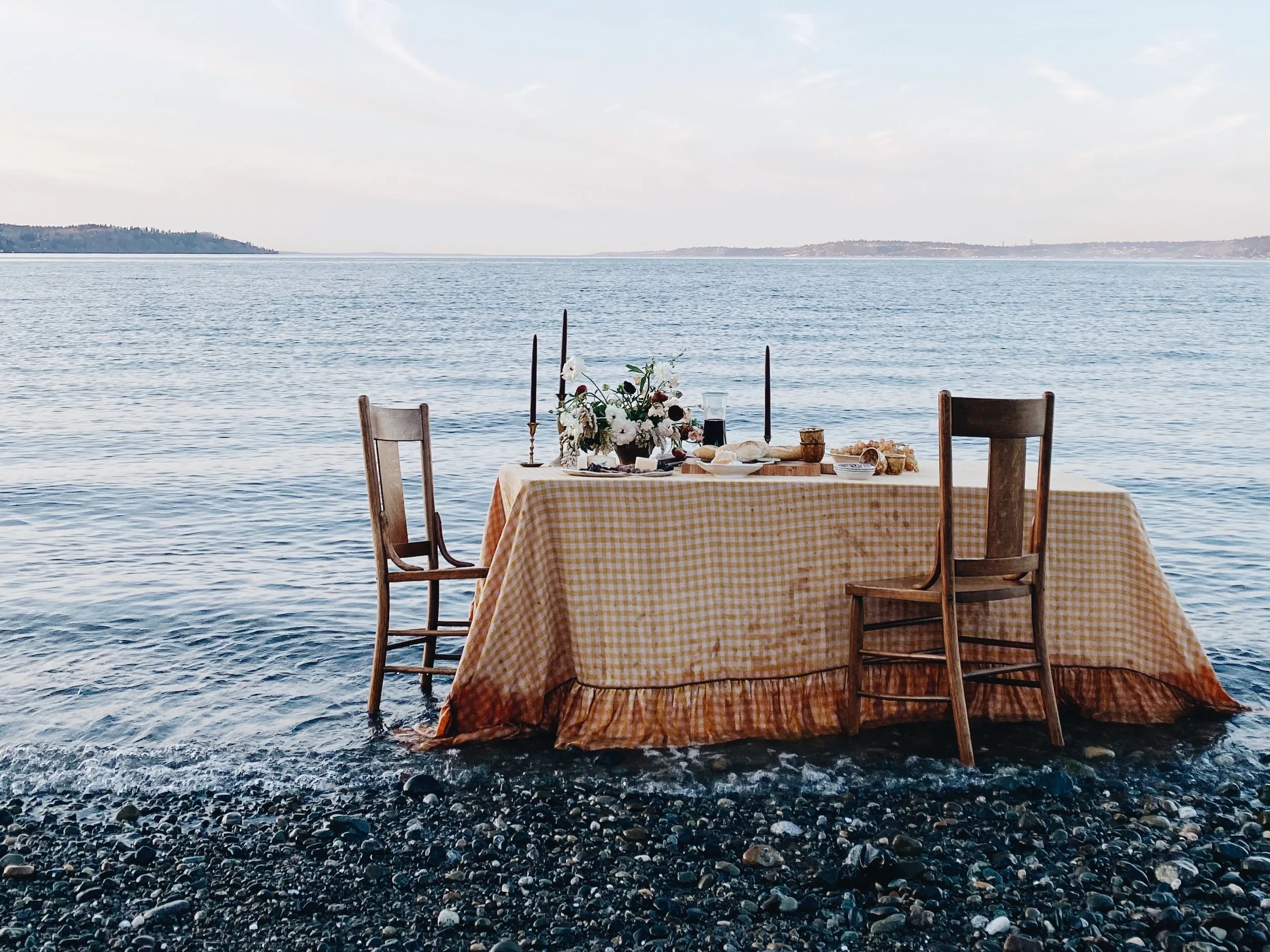 A dining table with a yellow checkered tablecloth set up on a rocky beach with two wooden chairs, a flower arrangement, candles, and food, overlooking a calm body of water under a cloudy sky.