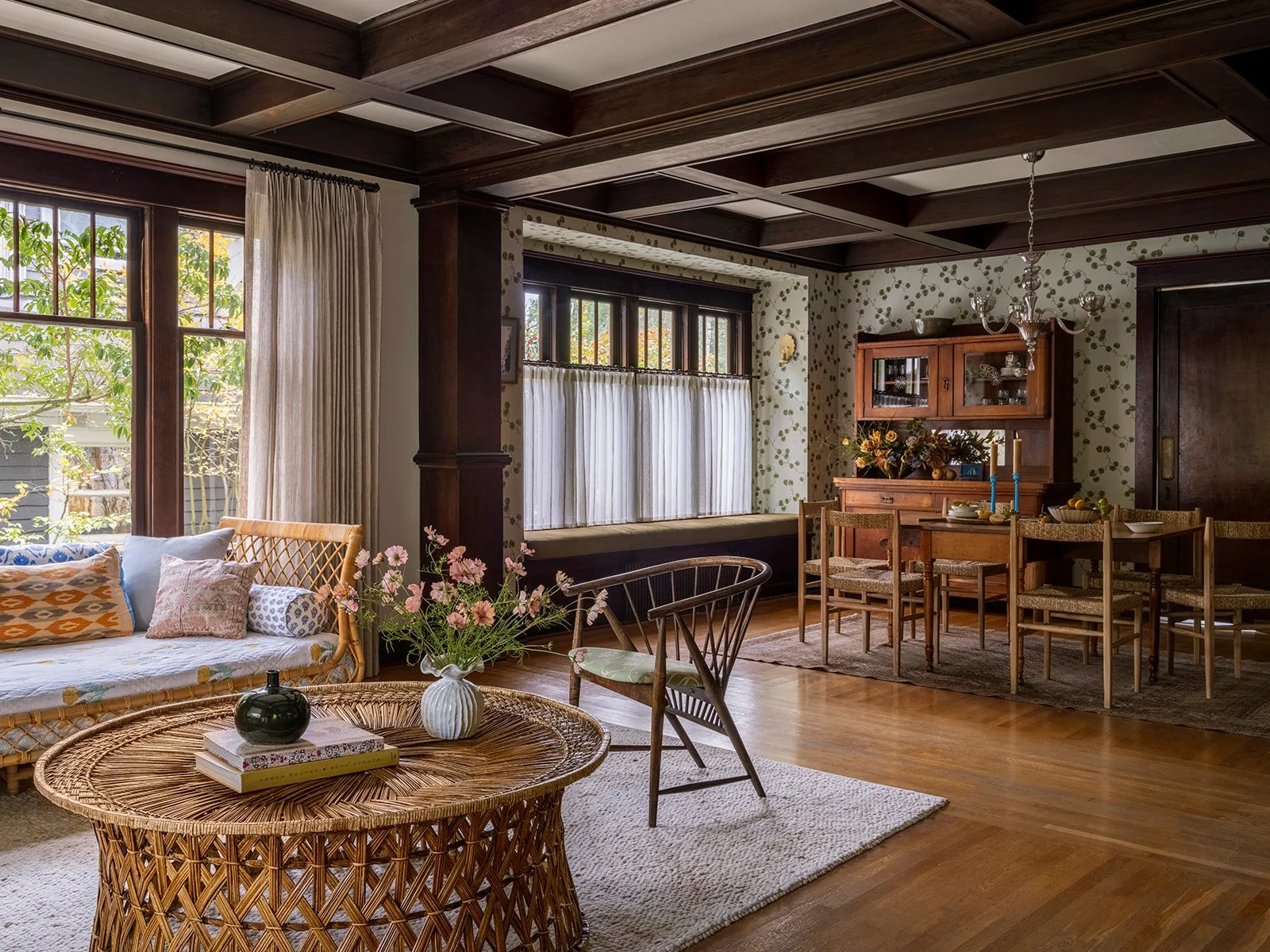 A living room with wooden beams on the ceiling, hardwood flooring, a wicker coffee table with books and a vase of flowers, a white couch with colorful pillows, a wooden dining table with chairs, and a wooden hutch on the wall, with large windows and curtains.