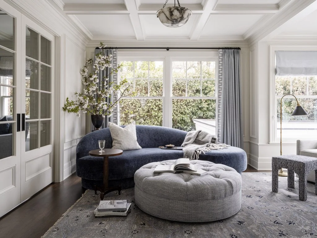 Bright living room with large windows, gray curtains, a blue velvet sofa with white pillows, a round gray ottoman with an open book, a small wooden side table with a glass, a floor lamp, and a patterned rug.