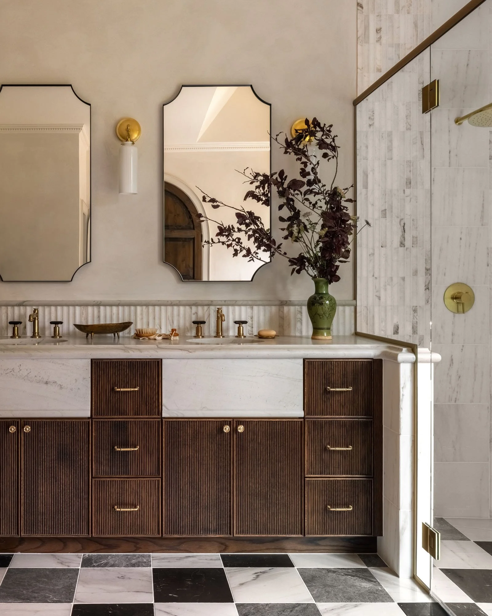 A bathroom vanity with a marble countertop, three mirrors, a green vase with dark branches, and various small decorative items, all reflected in the mirrors. The lower part of the vanity is made of dark wood with vertical slats and gold handles. There is a black and white checkered floor.