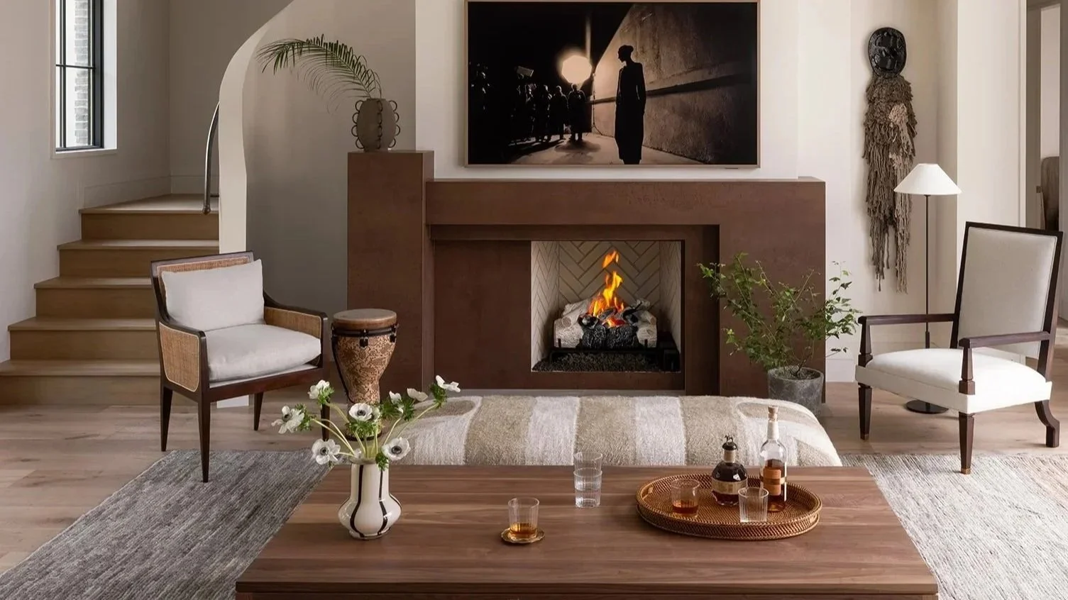 A cozy living room with a modern fireplace, two armchairs, a coffee table with drinks, a fireplace, a large painting, a small plant, and decorative objects.