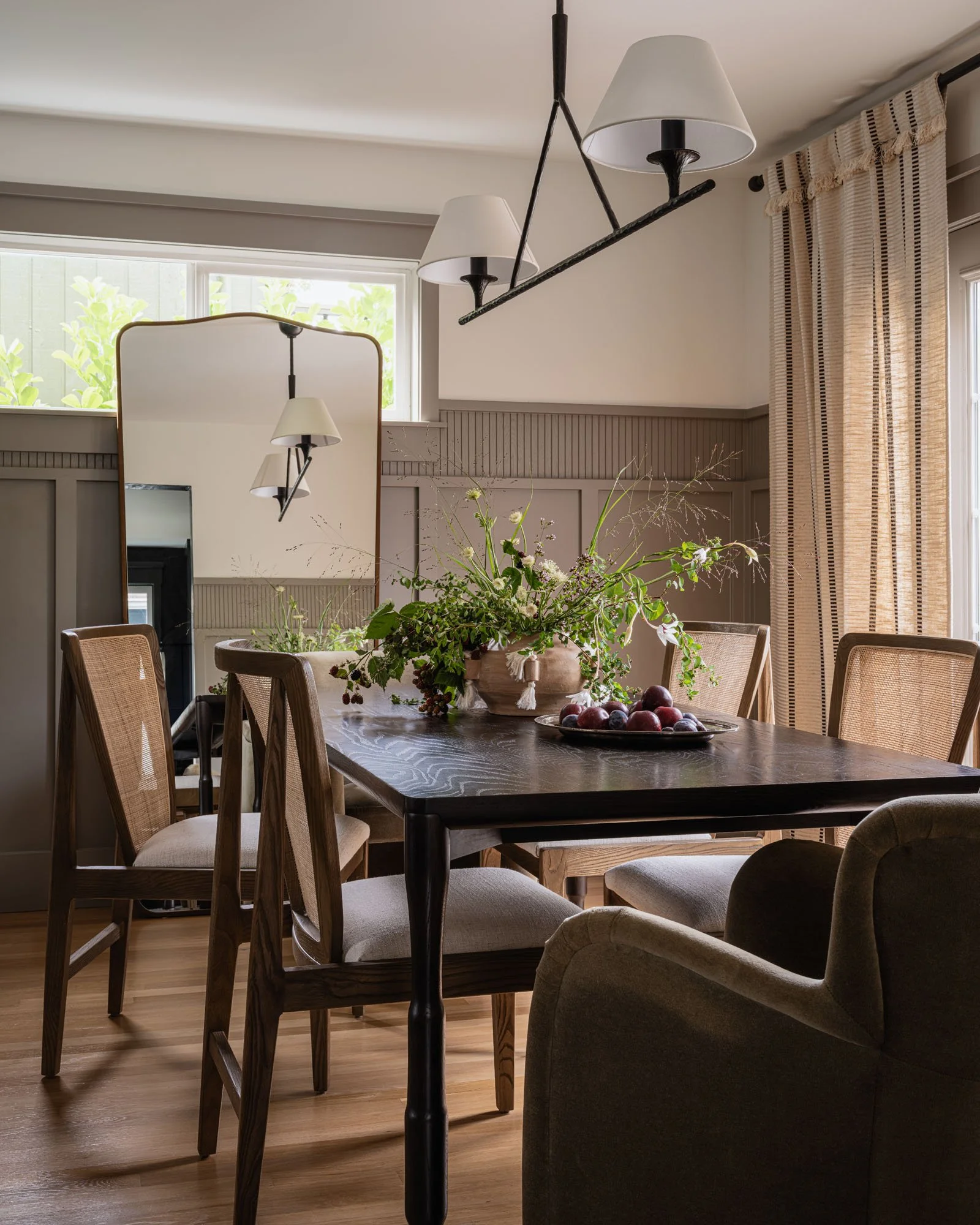 Dining room with a dark wooden table, six chairs, a large plant centerpiece, a mirror, and a modern light fixture with three lampshades.