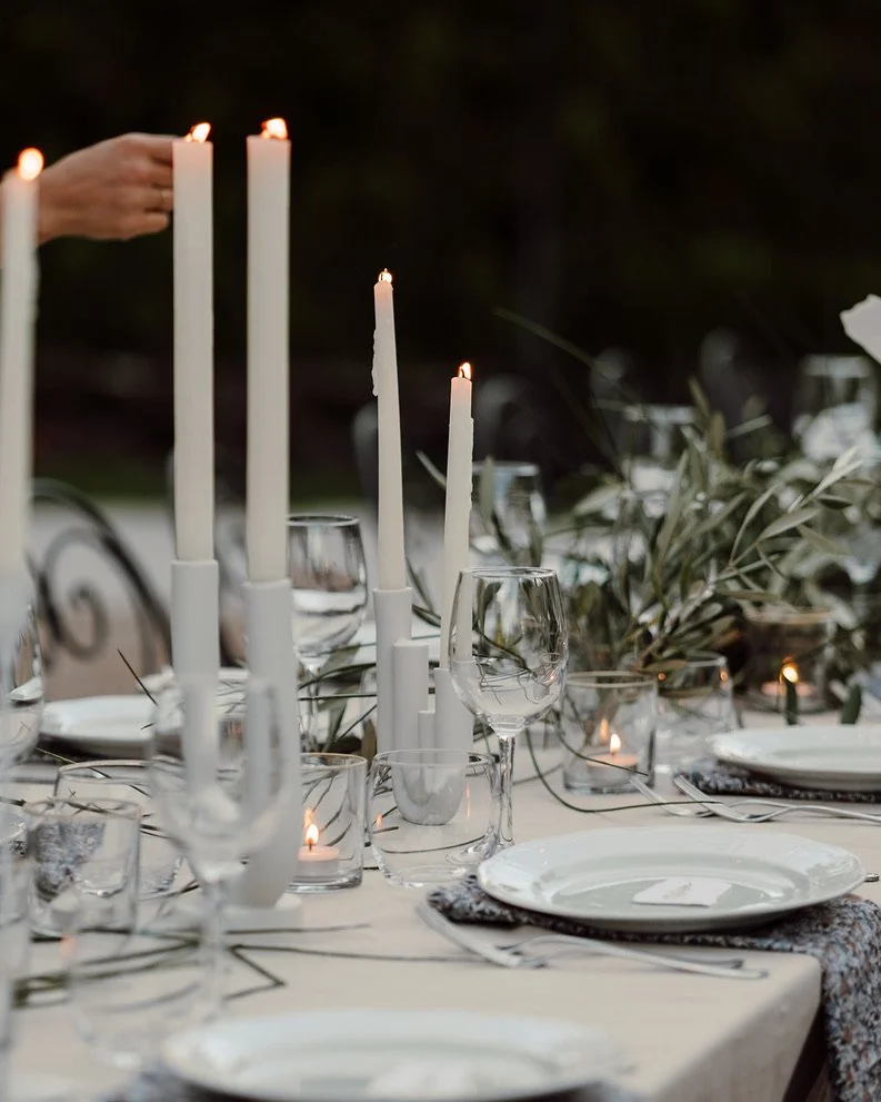 Elegant outdoor dining table decorated with white candles in holders, wine glasses, and greenery accents, set for an evening event.