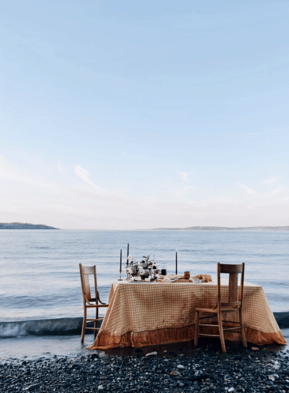 A dining table set up on a rocky beach by the water with a tablecloth, candles, and a vase of flowers.