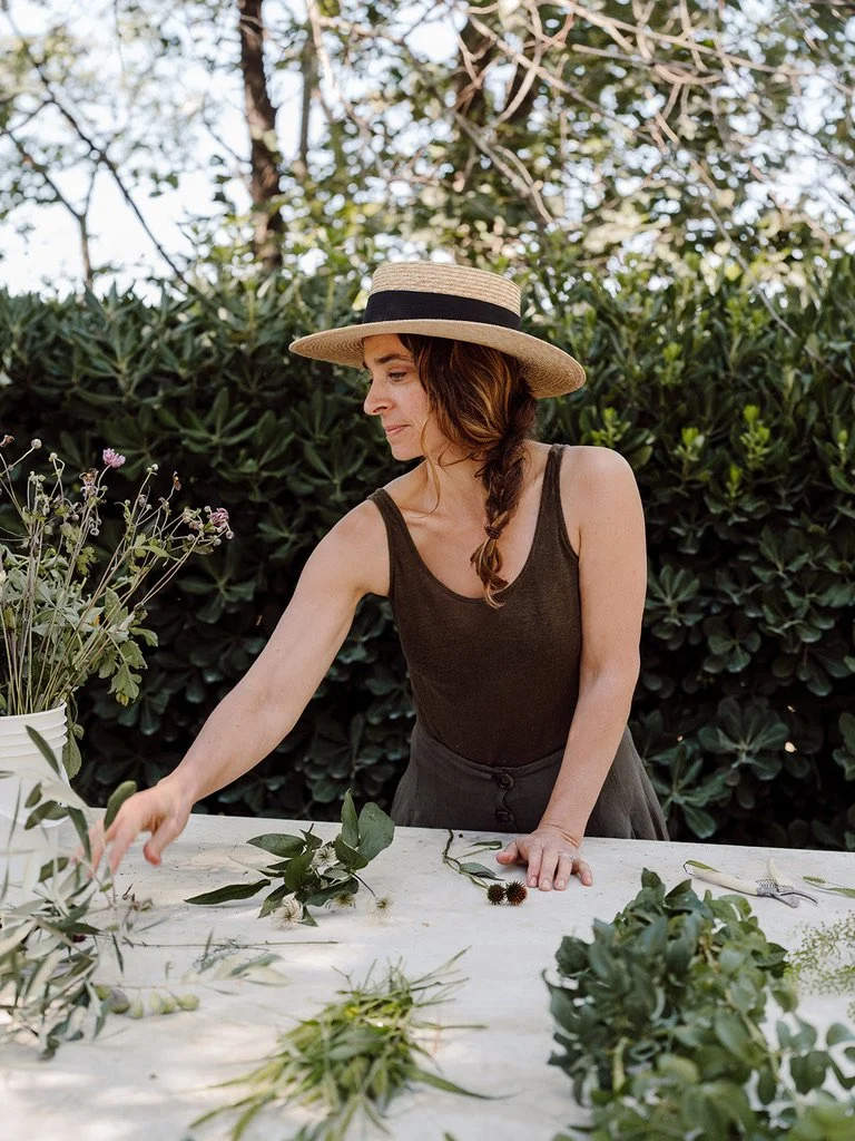 Woman wearing a straw hat with a black band, arranging flowers and greenery on a table outdoors.